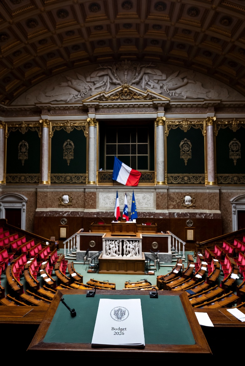 Style photojournalistique, salle de l'Assemblée nationale française baignée d'une lumière verdâtre, podium vide avec un dossier « Budget 2026 », drapeaux français et européen se confondant en arrière-plan, symbole de l'austérité financière sous contrôle européen, éclairage réaliste, 